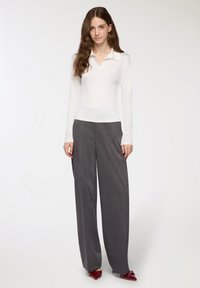 Polo bianco a maniche lunghe con colletto, abbinato a pantaloni grigi a gamba larga e scarpe con tacco rosse. Texture liscia e design aderente.