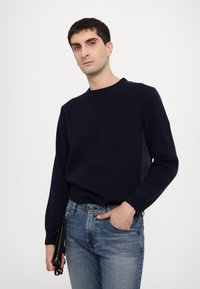 Pull en maille bleu marine avec un col rond, des poignets côtelés et une finition texturée, associé à un jean en denim bleu clair et un accessoire noir.