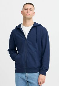 Marineblauwe ritshoodie gemaakt van een zachte stof, met een capuchon, trekkoorden, voorzakken en geribde manchetten, gecombineerd met lichtblauwe jeans.