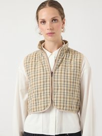 Mlada ženska, ki nosi bež in sivo karo jakno s quiltingom preko bele srajce z gumbi ter srebrne obročaste uhane.