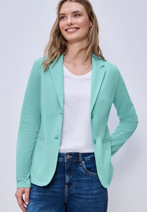 Lachende vrouw met een mintgroene blazer, wit overhemd en blauwe spijkerbroek, staand met één hand achter haar rug.