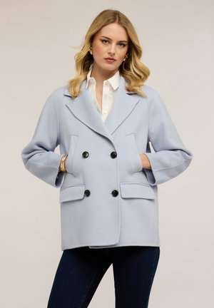 Blonde vrouw met lichtblauwe dubbelgeknoopte jas met zwarte knopen, wit overhemd, donkere broek, gouden creolen en armbanden, handen in de zakken.