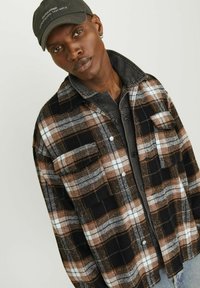 Black and brown checkered flannel jacket with white accents, featuring two chest pockets and a snap button closure, layered over a gray shirt.