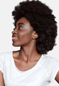 Boucles d'oreilles en or suspendues avec un design géométrique, portées par une personne avec des cheveux noirs texturés, portant un t-shirt blanc sur un fond neutre.