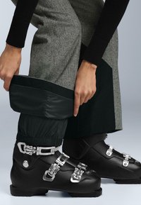 Fekete sícipő ezüst csatokkal, szürke halszálkás nadrággal és fekete elasztikus mandzsettával párosítva. A cipő karcsú dizájnnal rendelkezik.