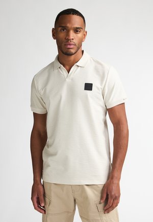 Beżowa koszulka polo z subtelnym, teksturowanym wzorem. Z przodu na lewej piersi znajduje się mała czarna naszywka z logo, posiada krótki rękaw.