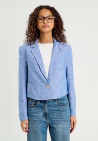 Blazer bleu cropped en tissu texturé, avec une fermeture à un bouton, des revers en pointe et des poches latérales, associé à un jean taille haute.