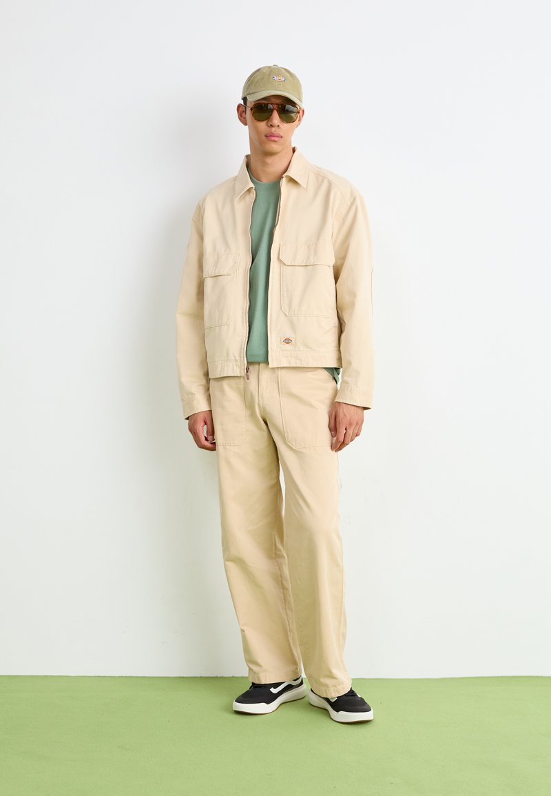 Veste en coton beige avec deux poches avant sur une chemise vert clair et un pantalon beige. Des baskets noir et blanc, une casquette verte et des lunettes de soleil complètent le look.