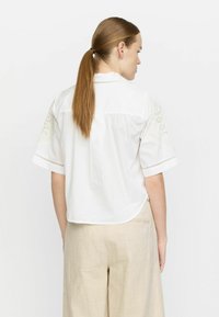 Weiße kurzärmelige Bluse mit gestickten Akzenten an den Schultern, mit abgerundetem Saum und lässiger Passform. Kombiniert mit beigen Leinenhosen.