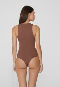 Mujer con cabello largo castaño que lleva un bodysuit sin mangas de color marrón, de pie de espaldas contra un fondo claro y sencillo.