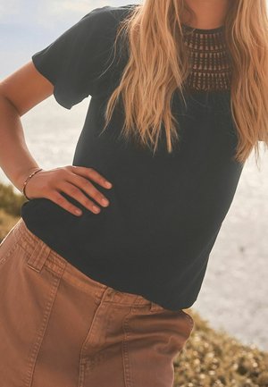 Musta lyhythihainen paita, jossa on teksturoitu ritiläkaulus, yhdistettynä beigeihin housuihin. Kangas näyttää pehmeältä ja siinä on rento malli.