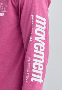 Camisa de algodón rosa de manga larga con texto blanco en la manga: "movimiento", "PERPETUO" y "PERSISTENTE". Textura suave, diseño casual.