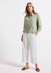 Camicia verde chiaro con maniche a tre quarti, caratterizzata da ricami a occhio sul fondo, abbinata a pantaloni bianchi a gamba larga e sandali marroni.