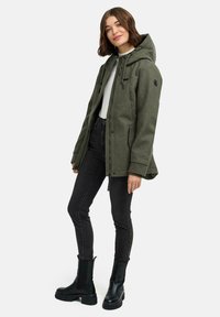 Femme debout portant une veste à capuche olive, un haut blanc, un jean skinny noir et des bottines noires sur un fond blanc.