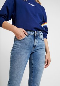 Sudadera azul marino con el logo de "Wrangler", combinada con jeans azules desgastados de cintura alta y bolsillos frontales. Material suave de algodón y mezclilla.