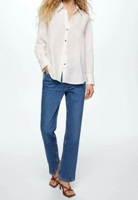 Kvinde iført en hvid langærmet skjorte med knapper, blå straight-leg jeans og brune strappy højhælede sandaler, poserer med den ene hånd i lommen.