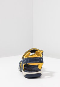 Bakåtsyn på en marinblå sandal med gula detaljer, utrustad med justerbara remmar och en strukturerad fotbädd. Vit sula med greppmönster.