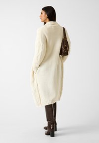 Lange roomkleurige gebreide cardigan met kabelpatroon op de mouwen, geribbelde manchetten en zoom; gecombineerd met bruine laarzen tot de knie met krokodillenprint.