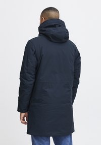 Parka bleu marine avec capuche, présentant une texture lisse, des manches longues et une longueur mi-cuisse. Design simple et fonctionnel.