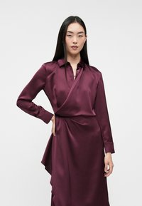 Burgundowa satynowa sukienka w stylu wrap z długimi rękawami, kołnierzykiem i asymetrycznym dołem. Posiada zapięcie na guziki oraz gładką, błyszczącą fakturę.