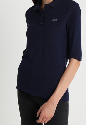 Kobieta ubrana w granatową koszulkę polo z krótkim rękawem z małym białym logo krokodyla na piersi oraz czarne spodnie.