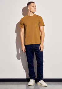 Hnědé tričko s kulatým výstřihem vyrobené z měkké tkaniny, v kombinaci s tmavě modrými džíny a bílými teniskami, stojící na světlejším pozadí.