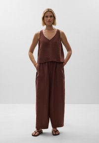 Rust-colored linen jumpsuit featuring a V-neck tank top with wide legs, front pockets, and a relaxed fit, paired with brown flip-flops.