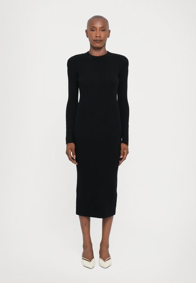 Schwarzes, geripptes Strickkleid mit langen Ärmeln, taillierter Silhouette, Rundhalsausschnitt und knielanger Länge, kombiniert mit weißen, spitzen Absatzschuhen.