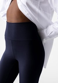 Legginsy w kolorze granatowym, o wysokim stanie, wykonane z gładkiego, elastycznego materiału. W połączeniu z luźną białą koszulą, odsłaniającą część pleców.