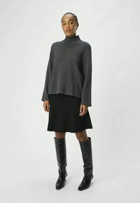 Woman standing wearing a dark gray turtleneck sweater, black knee-length skirt, and black knee-high boots against a plain light background.