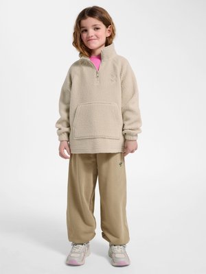 Ung pige stående iført beige fleecepullover, løse beige bukser med små grønne broderier, hvide sneakers og en lyserød skjorte under.