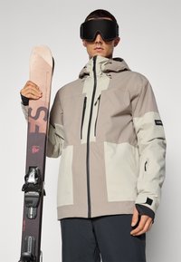 Ski jakna u svijetlo bež i krem boji, koja ima džepove s patentnim zatvaračem i kapuljaču. Osoba drži tamnu skijašku dasku s vezovima, nosi velike crne naočale.