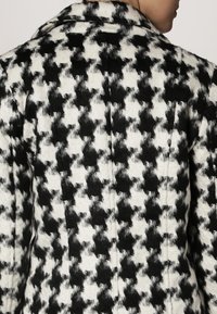 Schwarz-weiß kariert gemusterter Mantel mit einer strukturierten Stoffqualität, ausgestattet mit einem Reverskragen und sichtbaren Nähten auf der Rückseite.