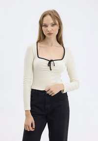 Maglia a maniche lunghe a coste di colore bianco sporco con scollatura quadrata, bordi neri e dettaglio con fiocco centrale, abbinata a jeans neri a vita alta.