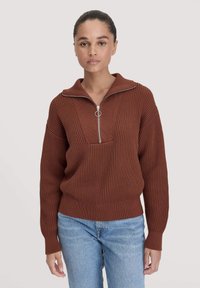 Bronzefarbenes Strickpullover mit halbem Reißverschluss-Kragen, gerippter Textur und breitem Schnitt, kombiniert mit hellblauen Jeans.