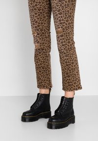 Svarta ankelstövlar med snörning och en tjock plattformssula, kombinerade med bruna jeans i leopardmönster med slitna detaljer och fransade fållar.