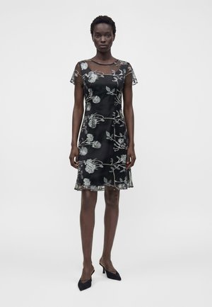 Femme portant une robe noire transparente avec une broderie florale argentée, manches courtes et talons noirs pointus, debout devant un fond uni.