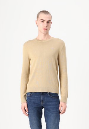 Suéter de punto beige con escote redondo, ribete en los bordes y mangas largas. Presenta un pequeño logotipo en el pecho. Combinado con unos vaqueros azules.