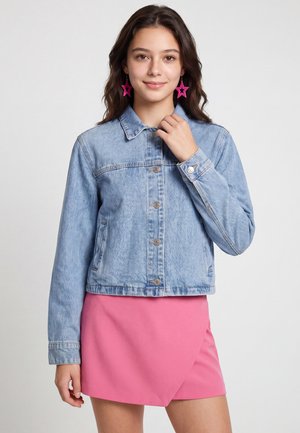 Ung kvinde med brunt hår iført blå denimjakke, lys pink nederdel og lyserøde stjerneformede øreringe, står mod en ensfarvet lys baggrund.