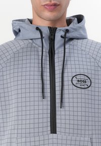 Sudadera con cremallera en gris que presenta un patrón en cuadrícula, acentos en negro, capucha con cordón y logo bordado en el pecho. Textura suave.