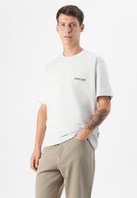 Hombre joven con cabello corto, vistiendo una camiseta gris claro con un logo, de pie de manera casual con una mano en el bolsillo y un tatuaje en el brazo.