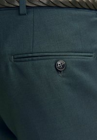 Mørkegrønne bukser med tekstureret stof, en baglomme med en sort knap og flad design.