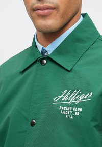 Πράσινο αδιάβροχο μπουφάν με γυαλιστερή υφή, που διαθέτει λευκό λογότυπο και κείμενο, καθώς και μαύρα κουμπιά στο μπροστινό μέρος. Ο ανοιχτό γαλάζιος γιακάς είναι ορατός.