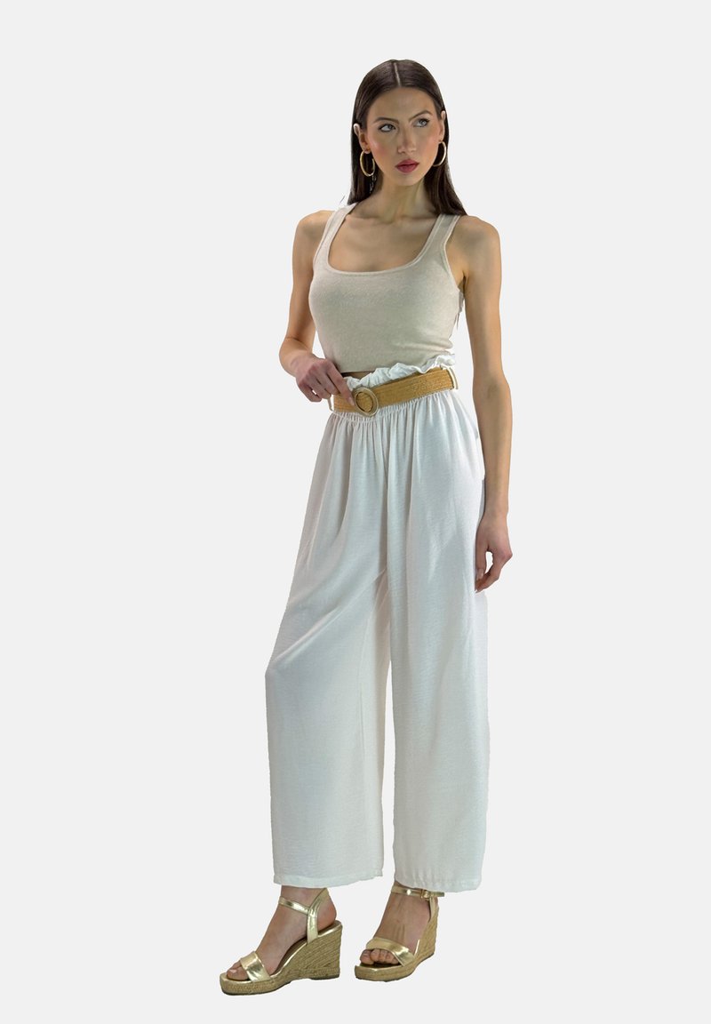 Beige tanktop med hvide, løse bukser og et struktureret, beige bælte. Gyldne wedgesandaler. Modellen står foran en neutral baggrund.