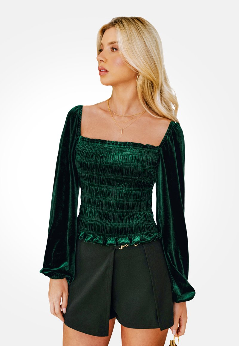 Top de terciopelo verde con escote cuadrado, textura fruncida, mangas largas y dobladillo con volantes; combinado con pantalones cortos negros con un detalle dorado.