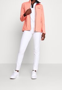 Veste légère corail avec fermeture éclair à l'avant, col noir et manches longues, associée à un pantalon slim blanc et des baskets blanches.