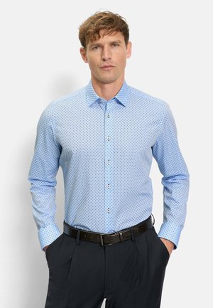 Mann trägt hellblau gemustertes Hemd mit Knöpfen, in dunkle Hose mit schwarzem Gürtel gesteckt, Hände in den Taschen, vor einfarbig weißem Hintergrund.