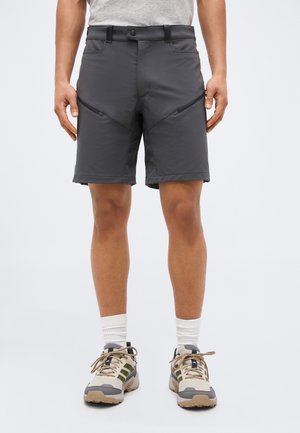Homme portant un short gris zippé, des chaussettes blanches et des chaussures de randonnée beige, debout devant un fond uni.
