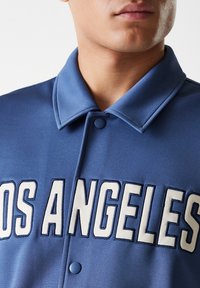 Blue button-up jacket with a smooth texture, featuring large white embroidered text "LOS ANGELES" on the front and a classic collar design.