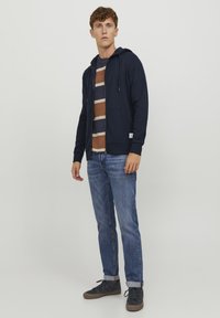 Mørkeblå zip-up hoodie med snor, båret over en stribet brun og beige skjorte, parret med lyseblå jeans og mørke sneakers.